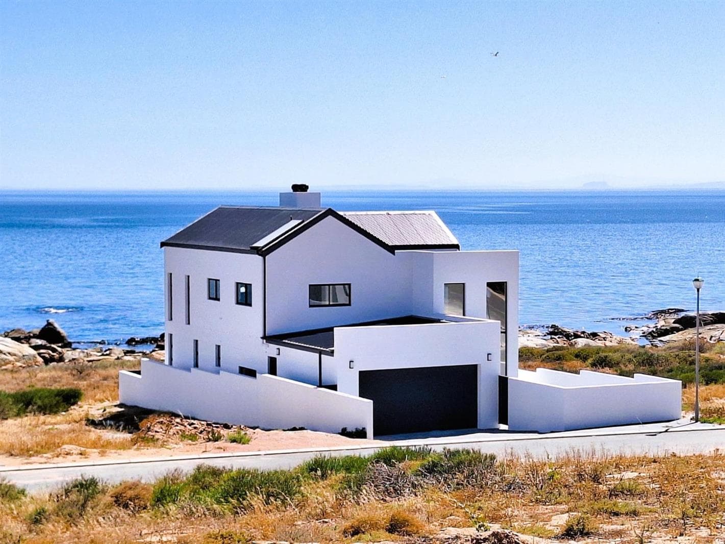 Modern white house with black roof overlooking the blue ocean from a rocky coastal landscape.