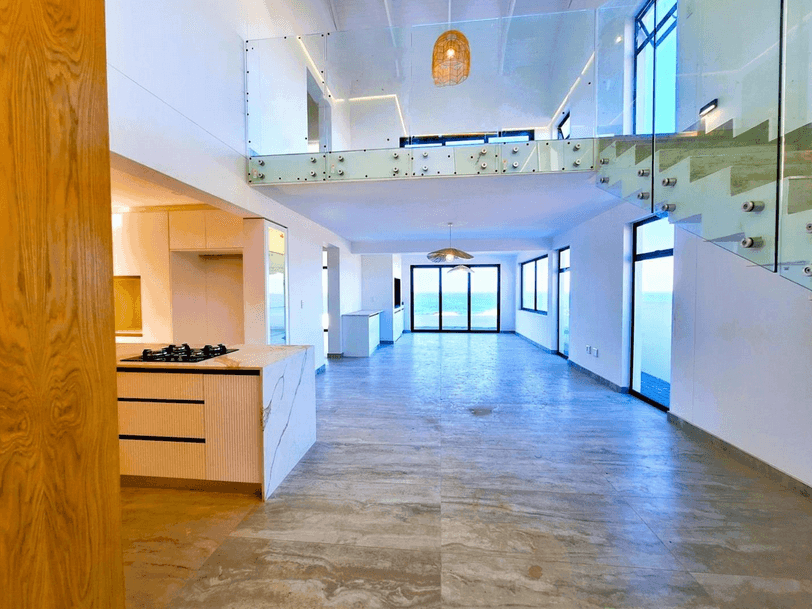 Spacious modern interior with kitchen island, glass-railed mezzanine, and large windows overlooking the ocean.