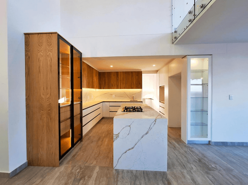 Modern kitchen featuring a marble island, wooden cabinetry, and tall illuminated glass display cases.
