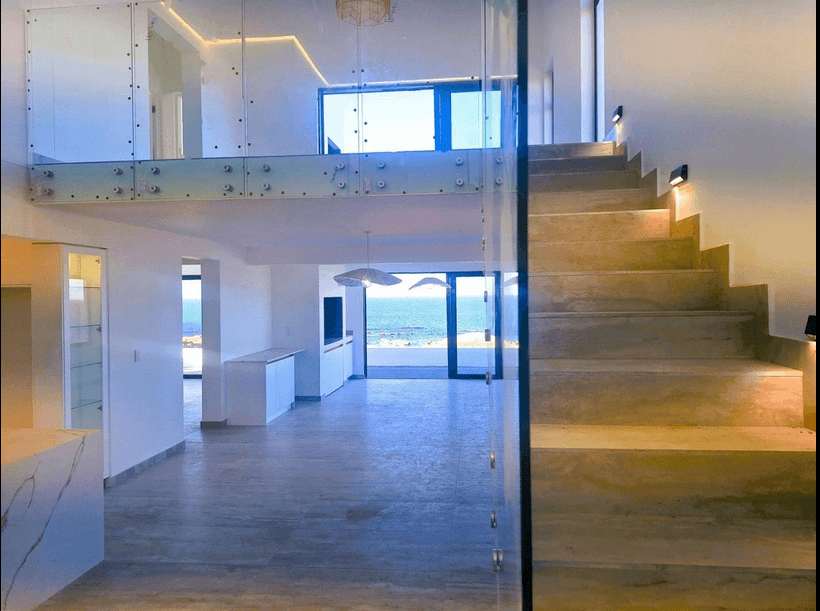 Modern minimalist interior with stone stairs, glass railings, and large windows overlooking the blue ocean.